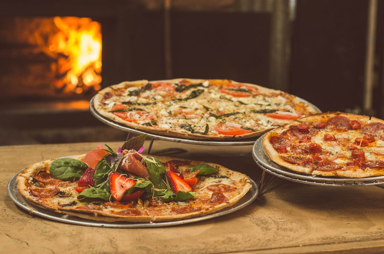 home-hero Trio of gourmet pizzas with unique toppings near a wood-fired oven, ideal for food enthusiasts.
